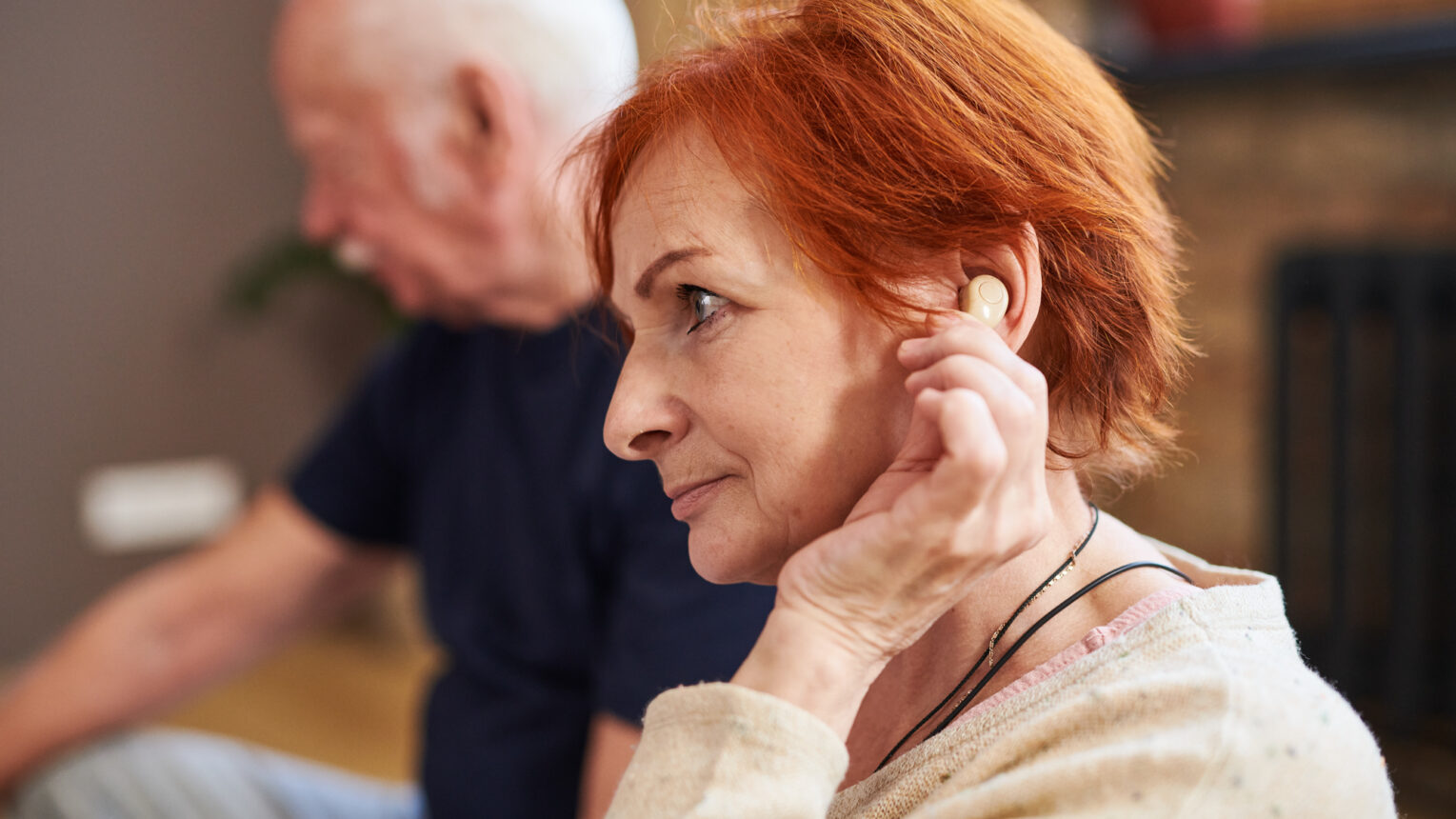 Bild - Sidovy av äldre kvinna med kort rött hår som använder sin hörapparat när hon sitter med andra människor i yogaklass